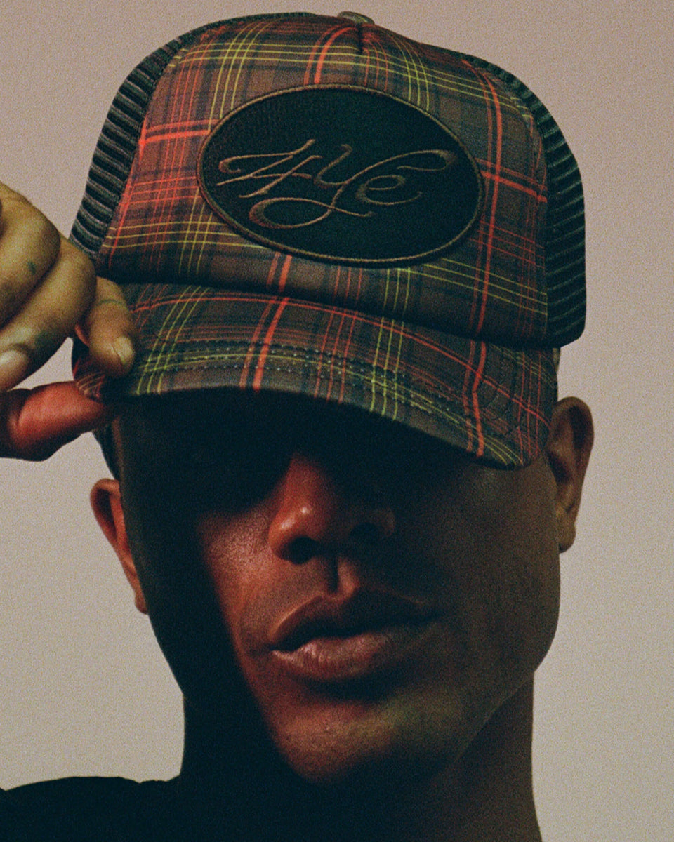 Model wearing a plaid trucker hat with a brand logo on a neutral background