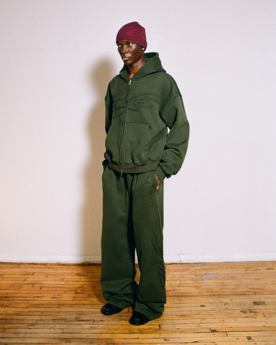 Model wearing dark green tracksuit and signature ribbed beanie plum/red standing against a white wall.