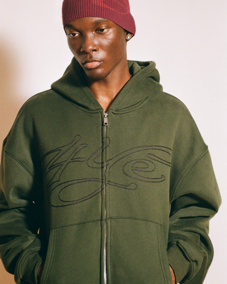 Model wearing a green zip up hoodie with black signature logo, standing against a neutral background