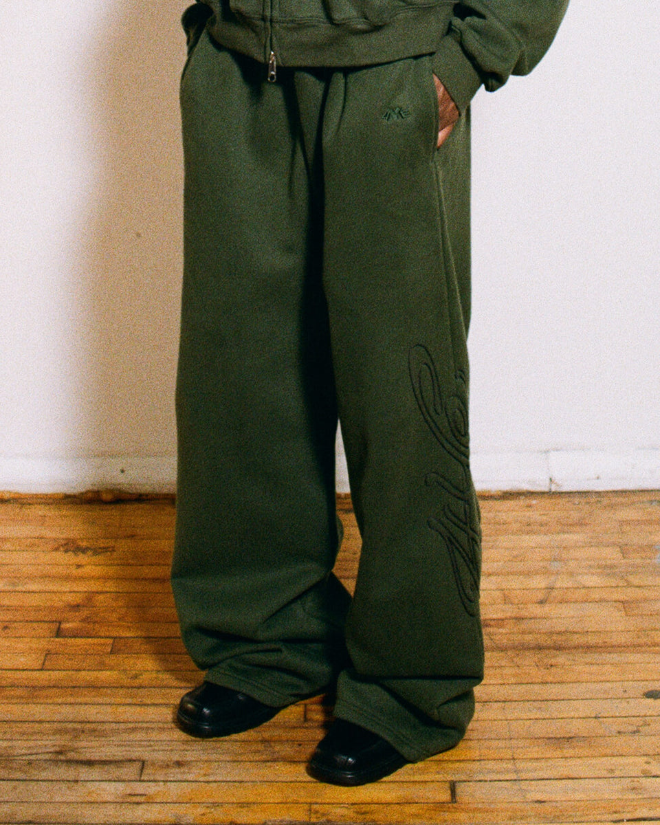 Model wearing dark green pants standing on a wooden floor with a white wall background