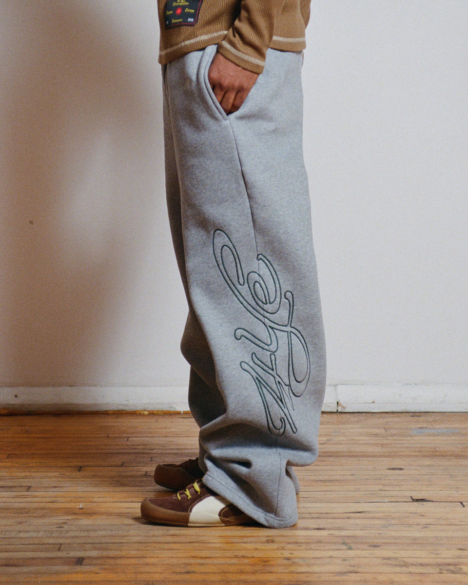 model wearing grey sweatpants with green logo, standing on a wooden floor.