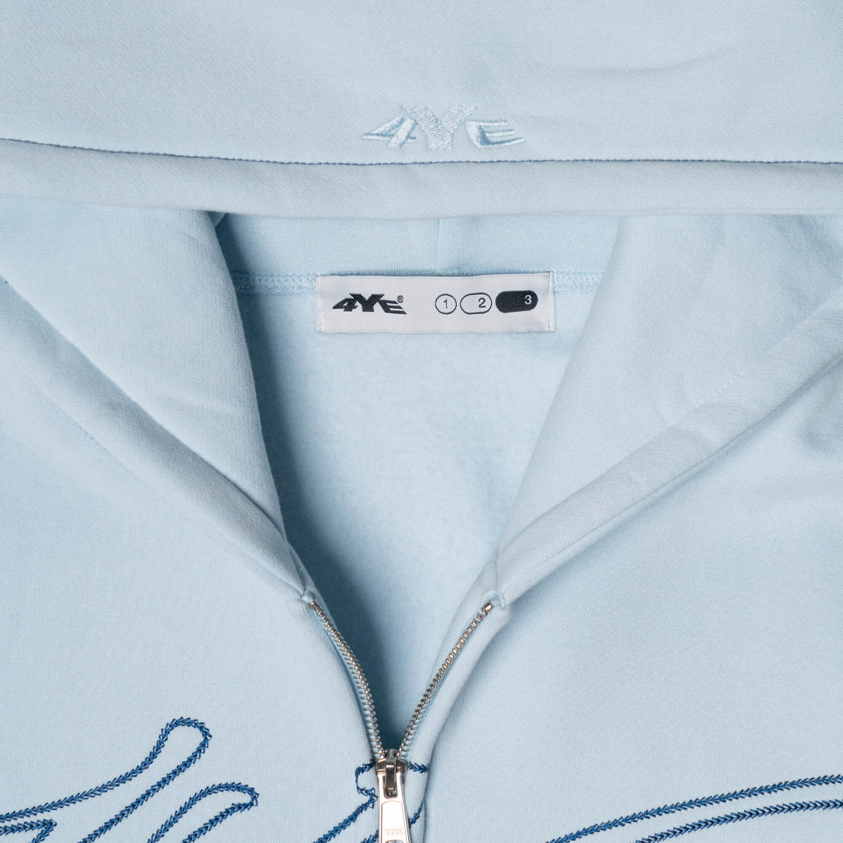 Close-up view of the 4YE Signature Logo Zip Hoodie in Light Blue. The hoodie features 325 GSM brushed medium weight fleece offering ultimate comfort and warmth, including high-quality chain stitch embroidery and a 2-way YKK zipper, showcasing a trendy cropped and boxy fit.